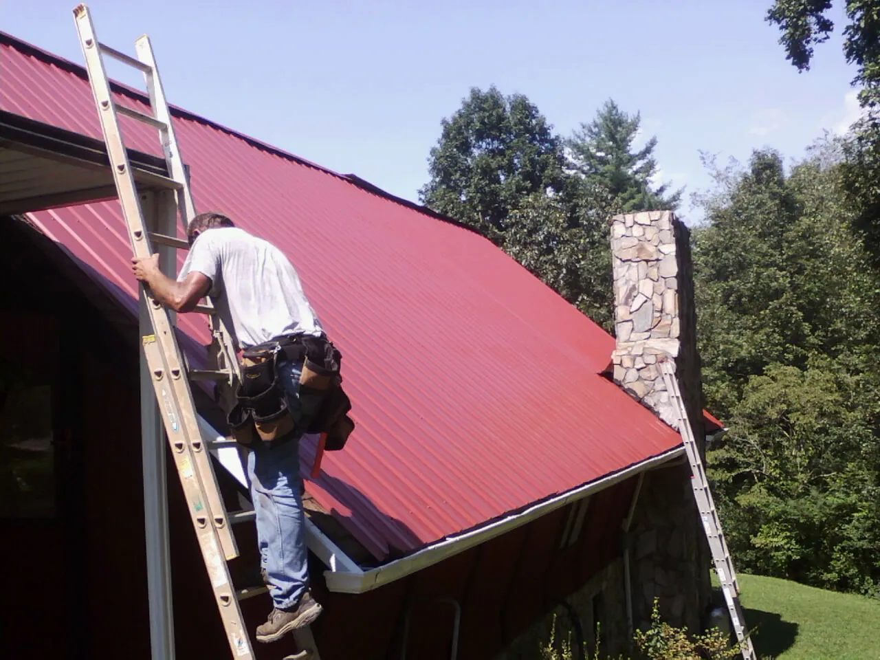 Roofer on a ladder beside a red metal roof and stone chimney — Soffit & Fascia Repair by Roofing Brothers in Highland Lakes